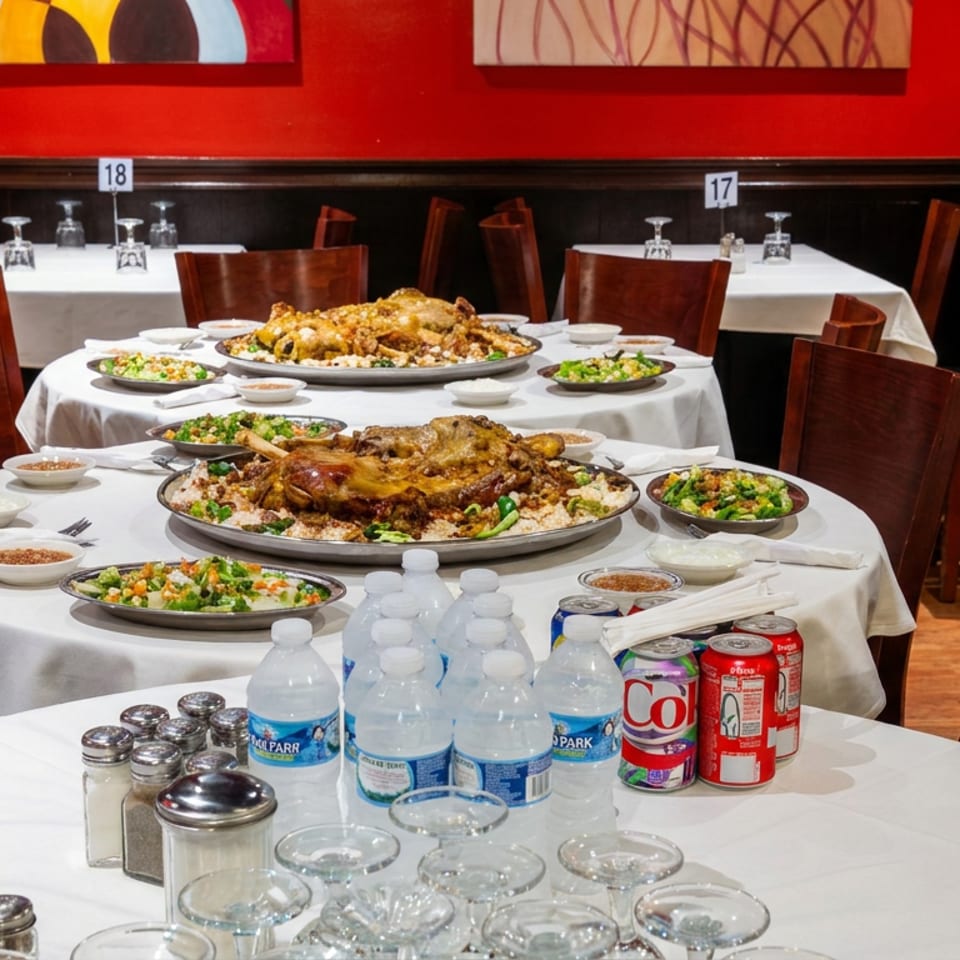 Restaurant dining room with red walls, white tablecloths, plated shared dishes, bottled water, soda cans, and empty chairs.