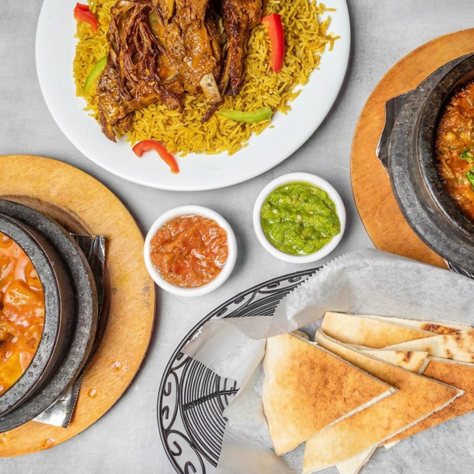 Overhead spread of spiced rice with grilled meat, two curries, naan wedges, and bowls of chutney on a gray table.