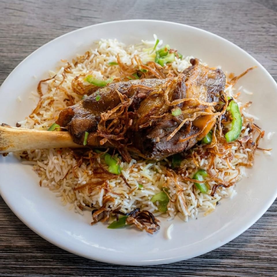 Plate of spiced rice topped with a roasted meat shank, fried onions, and green pepper garnishes on a wooden table.