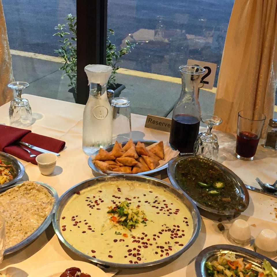 Middle Eastern restaurant table covered with hummus, dips, salad, fried pastries, drinks, and shared dishes beside a window.