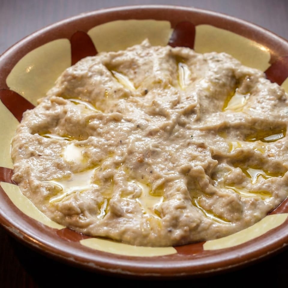 Creamy beige dip drizzled with olive oil sits in a patterned ceramic bowl on a dark wooden table.