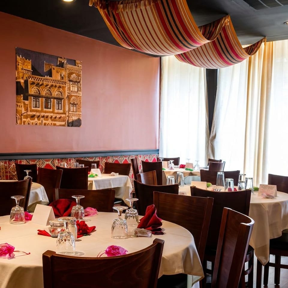 Warmly lit restaurant dining room with white tablecloths, red napkins, patterned textiles, artwork, and large windows.