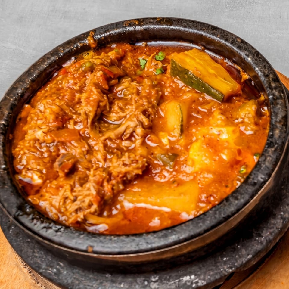 Rich red stew with shredded meat, potatoes, and zucchini served in a black stone bowl with flatbread.