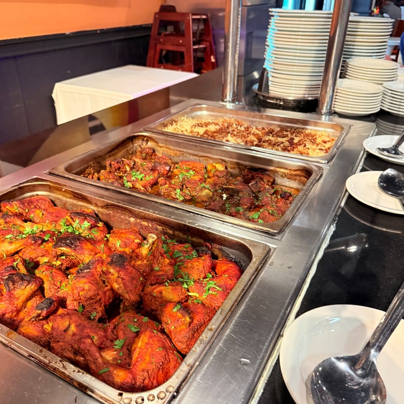 Middle-Eastern buffet with trays of chicken, stew, and rice, alongside stacks of white plates.