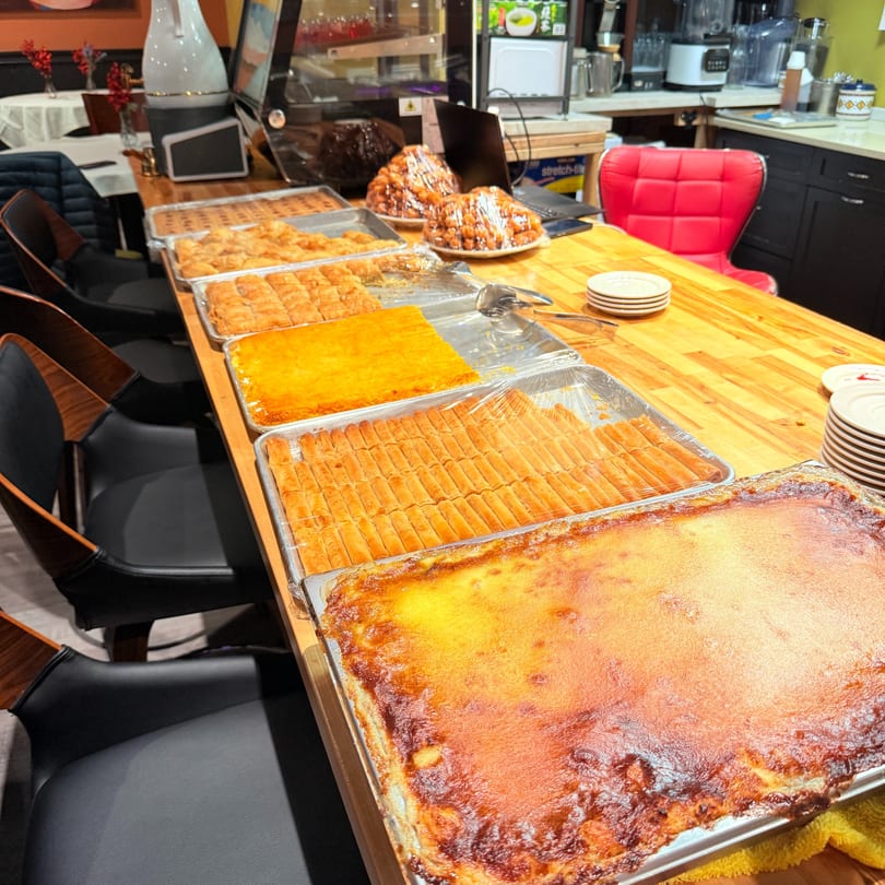 A long wooden counter displays trays of Middle Eastern desserts like baklava and a large baked custard.